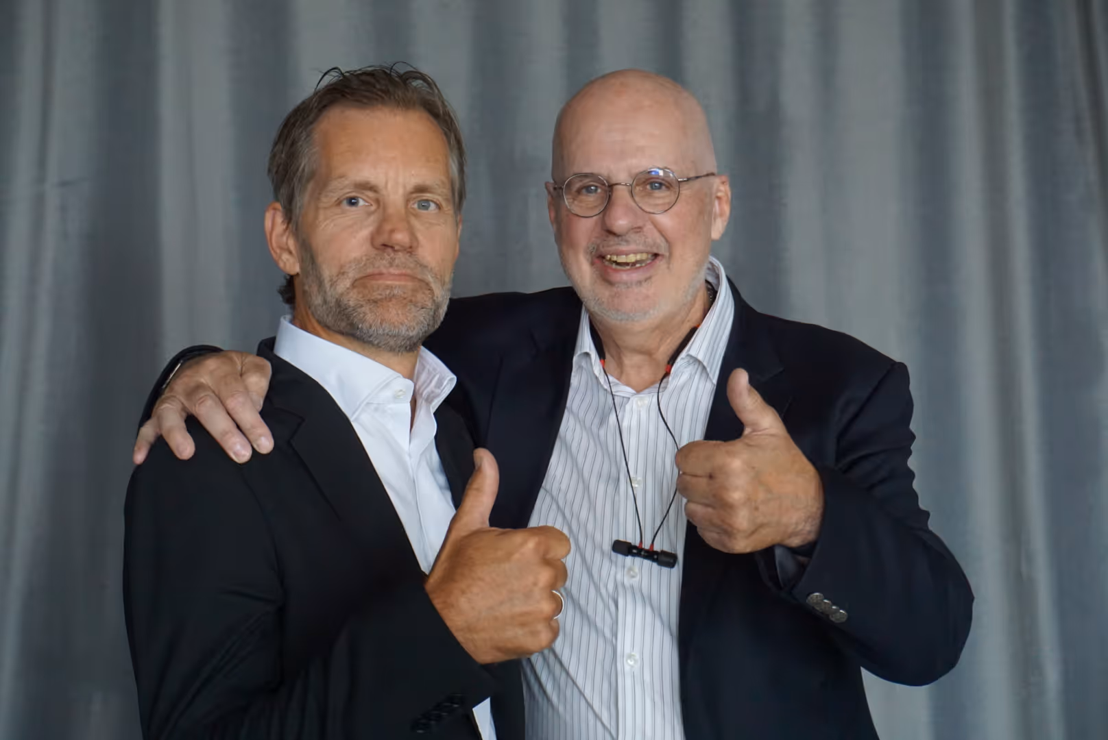 Två män i kostym poserar tillsammans mot en grå bakgrund, båda visar tumme upp och ler.