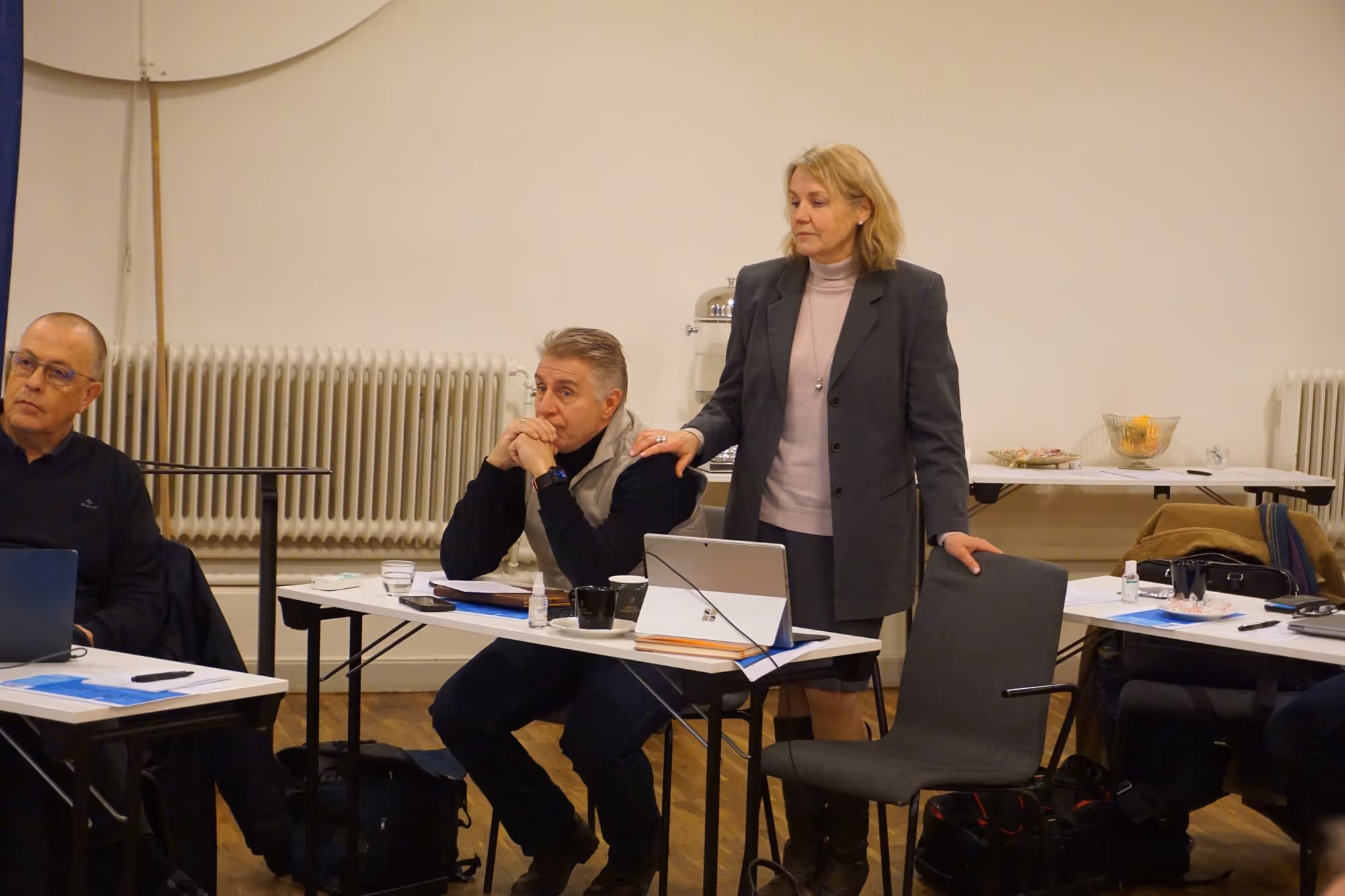 Kvinna i grå kavaj står bredvid sitt bord med en man som sitter och lutar huvudet på händerna i ett mötesrum.
