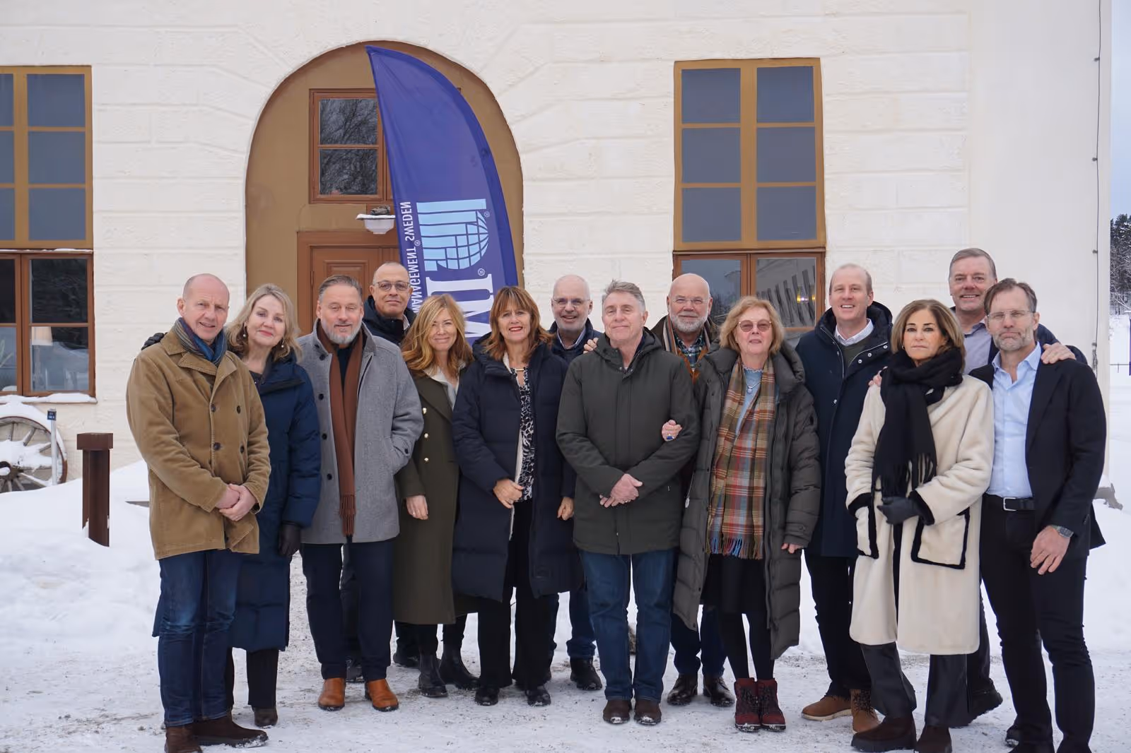 En grupp med 14 vuxna personer i vinterkläder står tillsammans framför en snötäckt byggnad med två stora fönster och en blå flagga.